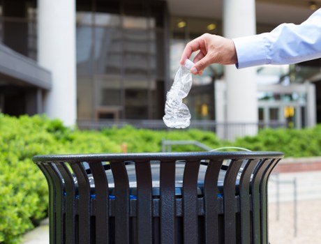 A person's hand, wearing a light blue long-sleeved shirt, is shown dropping a clear plastic water bottle into a black, slatted outdoor rubbish bin situated on a paved area. Behind the bin, there are blurred elements of a modern urban environment, including glass windows and metal structures, indicating a city or town setting. The water bottle is partially crushed, with a transparent texture that reflects natural daylight. The surrounding landscape includes well-maintained green bushes, and the scene is brightly lit with clear, natural light. This image visually supports rubbish removal services in the Kings Cross area, focusing on waste disposal and recycling. The context suggests an outdoor public or commercial space, aligning with professional waste management and rubbish collection offerings by Kings Cross House Clearance.