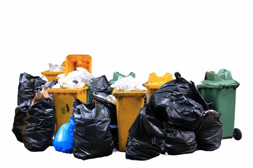 A collection of rubbish bins and bags arranged outdoors on a paved surface, situated against a plain white background. There are five wheeled bins in various colours including yellow, green, and black, with some lids open or partially open, revealing mixed waste such as paper, plastic, and miscellaneous refuse. Additionally, multiple large black trash bags are placed around the bins, some tied and bulging with contents, others loosely gathered. The scene appears to be a typical rubbish clearance setup, with waste materials awaiting collection, reflecting the type of clutter Kings Cross House Clearance handles in their services. The environment suggests an outdoor area adjacent to a property, possibly a driveway or communal space, prepared for rubbish removal by a professional team like Kings Cross House Clearance. The textures of the bins range from smooth plastic to the crinkled surface of the black rubbish bags, with the waste contents visible in some open lids, contributing to a realistic depiction of domestic or commercial waste awaiting disposal.