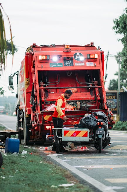 A sanitation worker, dressed in a yellow high-visibility vest with red accents, is unloading waste materials from the rear compartment of a large red rubbish collection truck parked on the side of a p